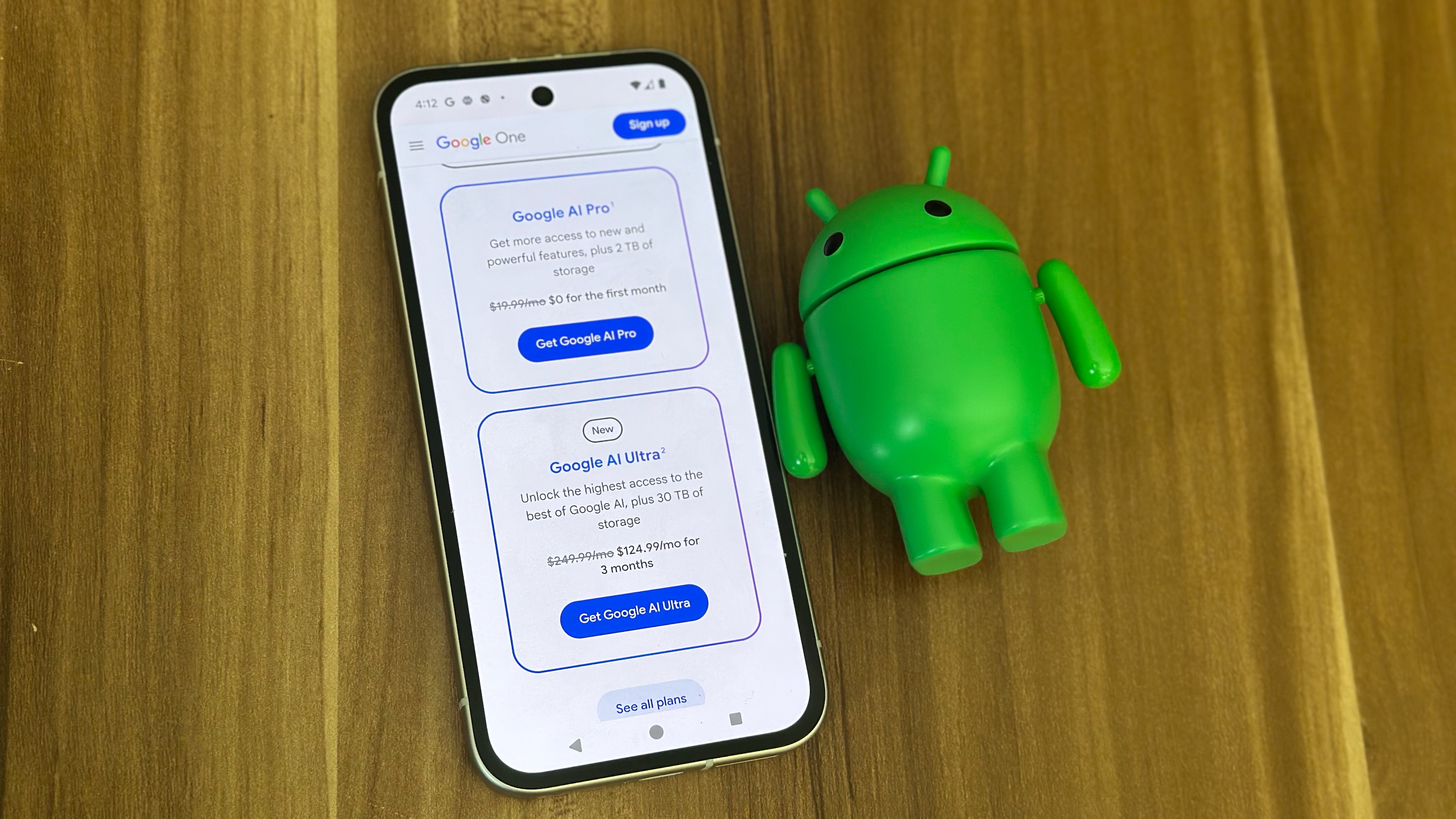 A Pixel 9a sitting next to an Android figurine showing the Google One AI Pro and AI Ultra plans.