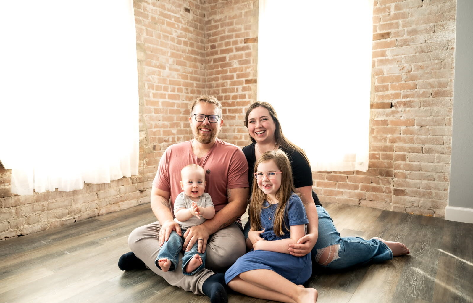 A mother and father sit on the floor in a well-lit room with their two young children in their laps for a studio portrait.