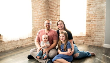 A mother and father sit on the floor in a well-lit room with their two young children in their laps for a studio portrait.