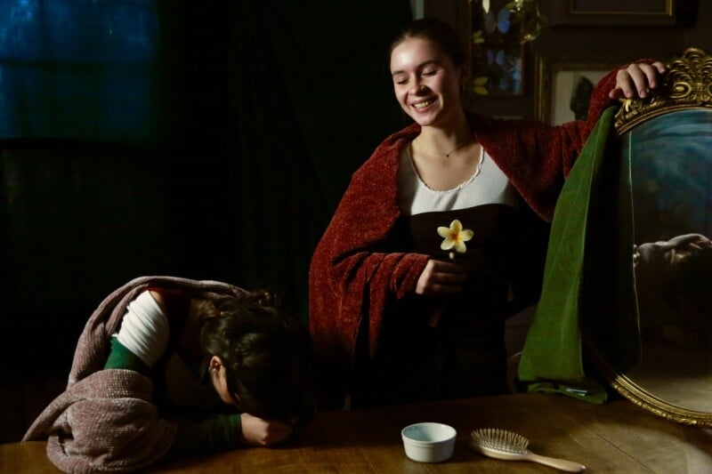 Two women in historical clothing are indoors; one is smiling and holding a flower and a mirror, while the other bows her head onto the table, partially covered by a blanket. A hairbrush and bowl sit on the table.