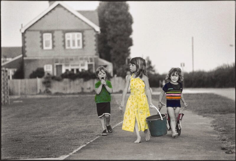 Three young children walk along a paved path in front of a house. The girl in the center wears a yellow dress and carries a green bucket, while the two boys beside her wear casual clothes and appear to be talking or eating.