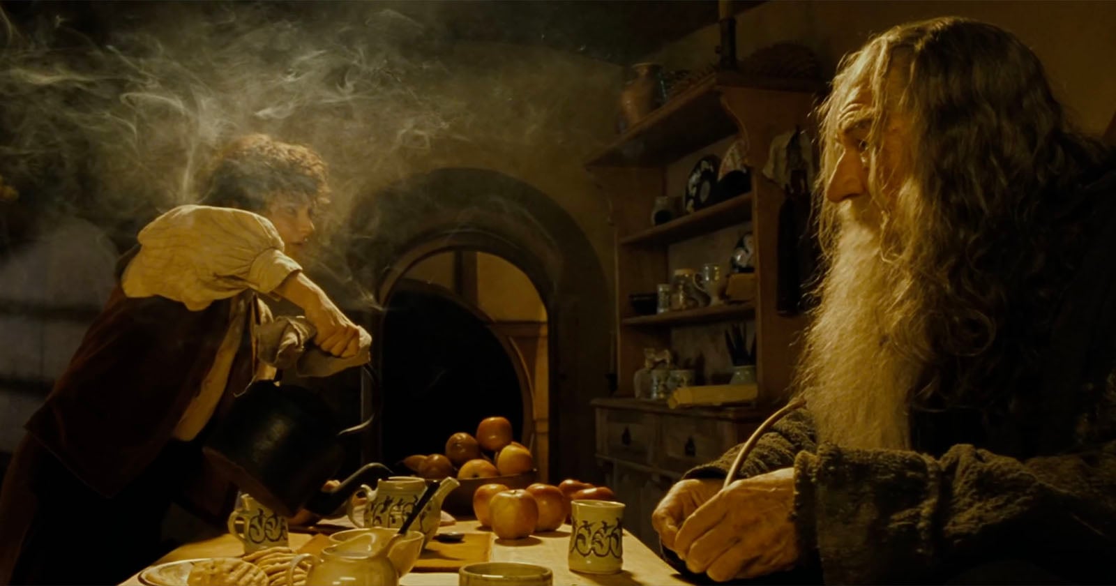 A young man pours tea from a kettle as an older bearded man sits at a wooden table with apples, mugs, and snacks in a cozy, warmly lit kitchen surrounded by shelves and dishes. Steam rises in the air.