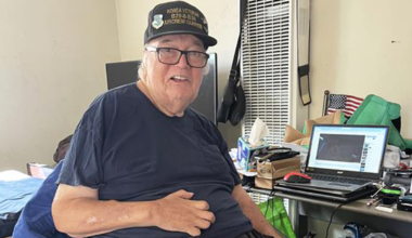 Elderly man wearing glasses and a "Korea Veteran" cap sits on a bed, smiling. A cluttered desk with a laptop and small American flag is beside him. He was able to return home due to Age-Friendly team.