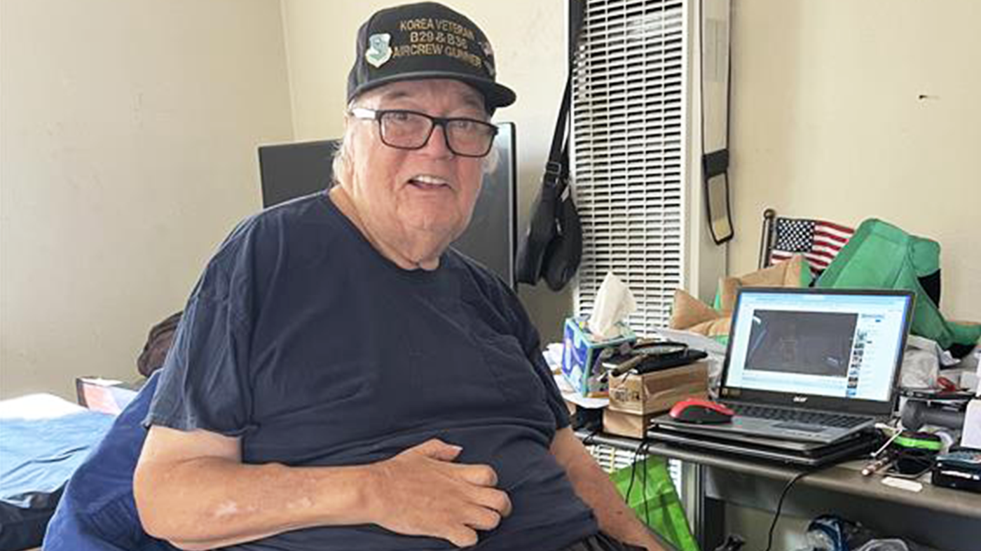 Elderly man wearing glasses and a "Korea Veteran" cap sits on a bed, smiling. A cluttered desk with a laptop and small American flag is beside him. He was able to return home due to Age-Friendly team.