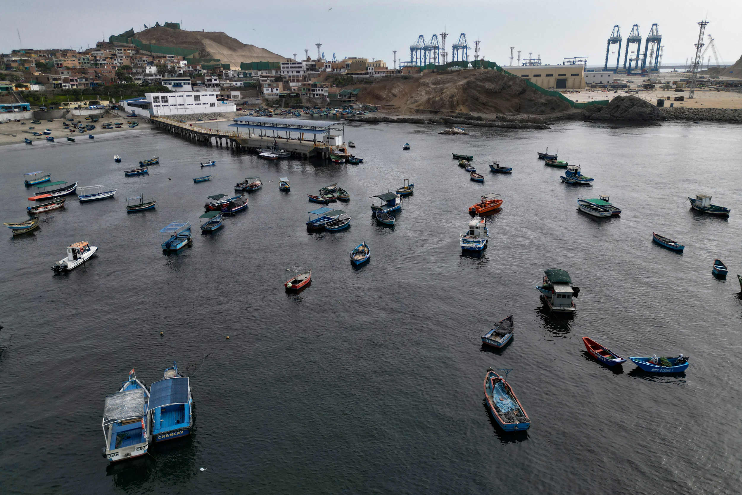 Fishing boats sit anchored in Chancay’s harbor with the new port’s cranes in the background. Credit: Cris Bouroncle/AFP via Getty Images