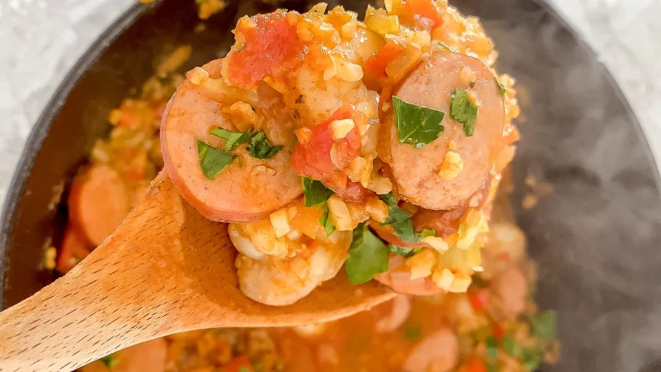 Close-up of a wooden spoon holding a serving of jambalaya with sliced sausage, shrimp, chopped vegetables, and fresh herbs over a skillet.