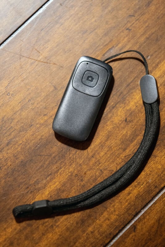 A small black electronic device with a single button and a wrist strap is lying on a wooden surface.