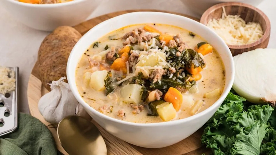A bowl of creamy soup with sausage, potatoes, kale, carrots, and beans, garnished with shredded cheese, surrounded by fresh greens, parmesan, potatoes, a garlic, an onion, and a golden spoon.