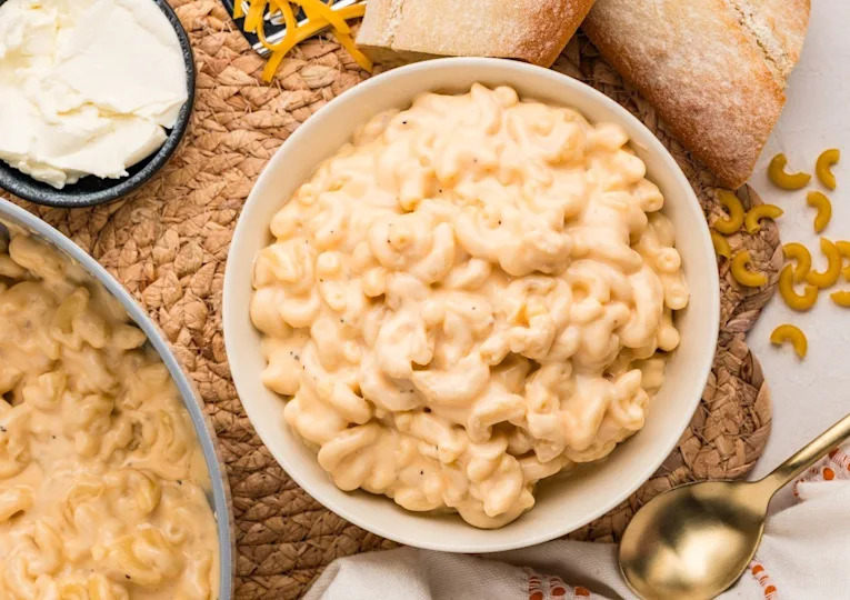 A bowl of creamy macaroni and cheese on a woven placemat, with a spoon, baguette slices, and a small dish of cheese nearby.