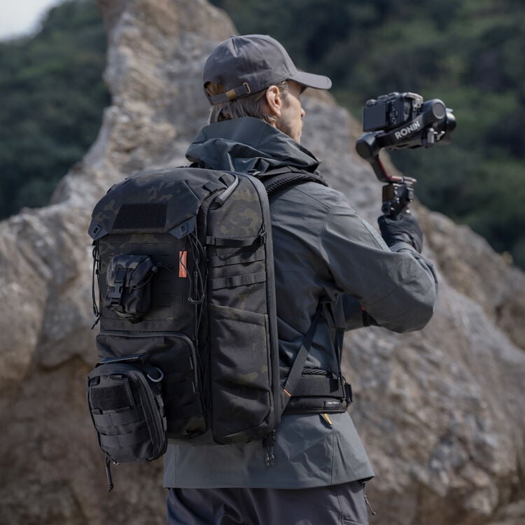 A person wearing a gray jacket, cap, and large black tactical backpack stands outdoors on rocky terrain, holding a camera mounted on a handheld gimbal, with trees visible in the background.