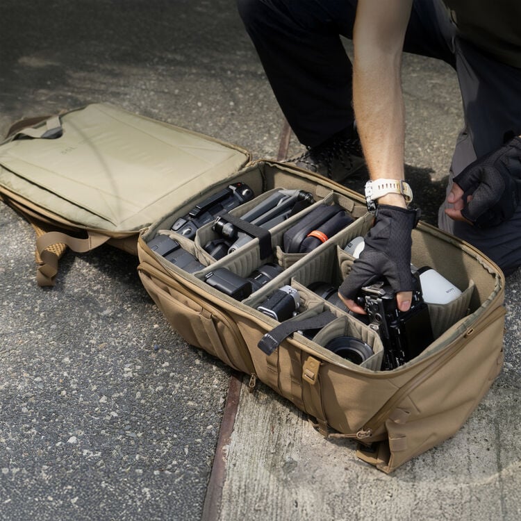 A person wearing fingerless gloves organizes camera equipment and accessories in a tan padded gear bag, which is open on the ground next to them.