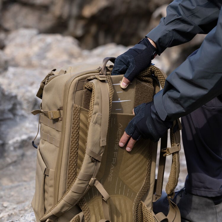 A person wearing black gloves and a gray jacket inserts a rectangular panel labeled "Aegis Frame" into the back compartment of a tan backpack on a rocky outdoor surface.