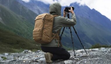 A person in a hooded jacket and tan backpack kneels on rocky ground, taking a photo with a camera on a tripod, with mountains and greenery in the background.