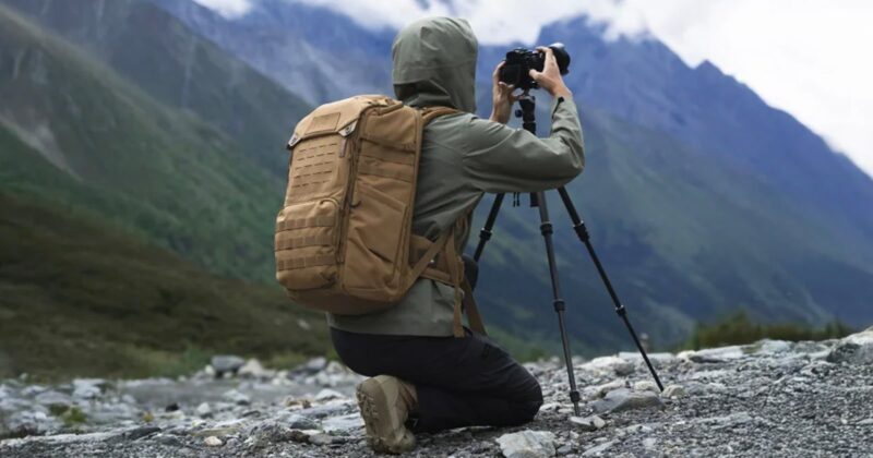 A person in a hooded jacket and tan backpack kneels on rocky ground, taking a photo with a camera on a tripod, with mountains and greenery in the background.