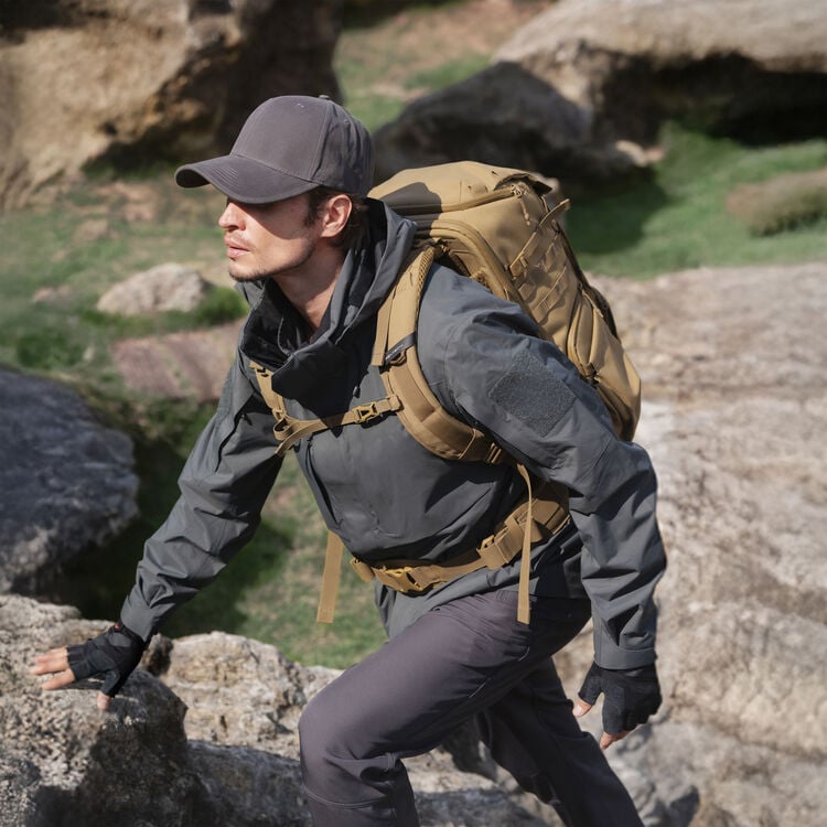 A man wearing a gray jacket, cap, gloves, and brown backpack is climbing over rocky terrain outdoors, looking ahead with focus. Green grass and large rocks are visible in the background.