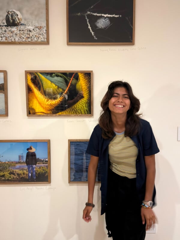 A smiling young person with medium-length dark hair stands in front of a wall displaying framed nature photos. They wear a striped shirt, dark open shirt, and bracelets, posing casually with one hand by their side.
