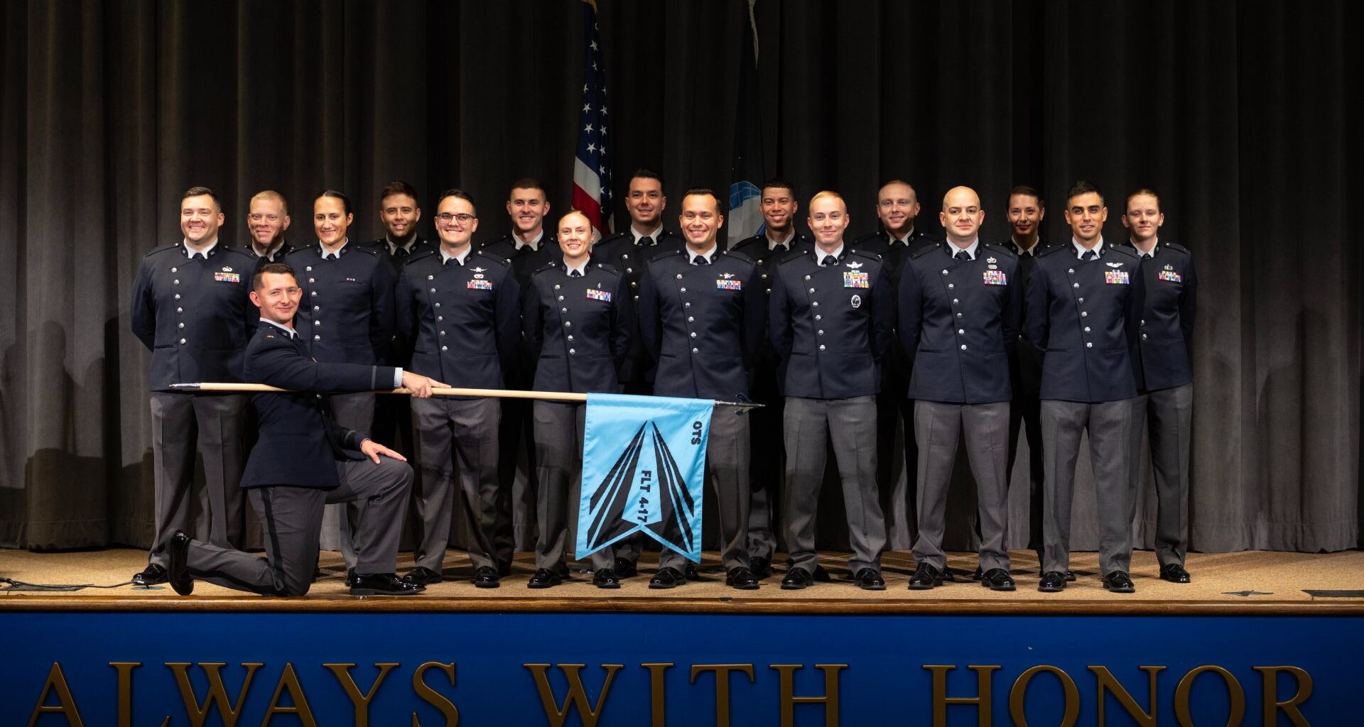 A group of people wearing dark blue shirts and black pants stand on stage with an American flag behind them and an officer kneeling down with a blue flag for the US Space Force placed in front of them.