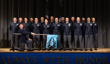 A group of people wearing dark blue shirts and black pants stand on stage with an American flag behind them and an officer kneeling down with a blue flag for the US Space Force placed in front of them.