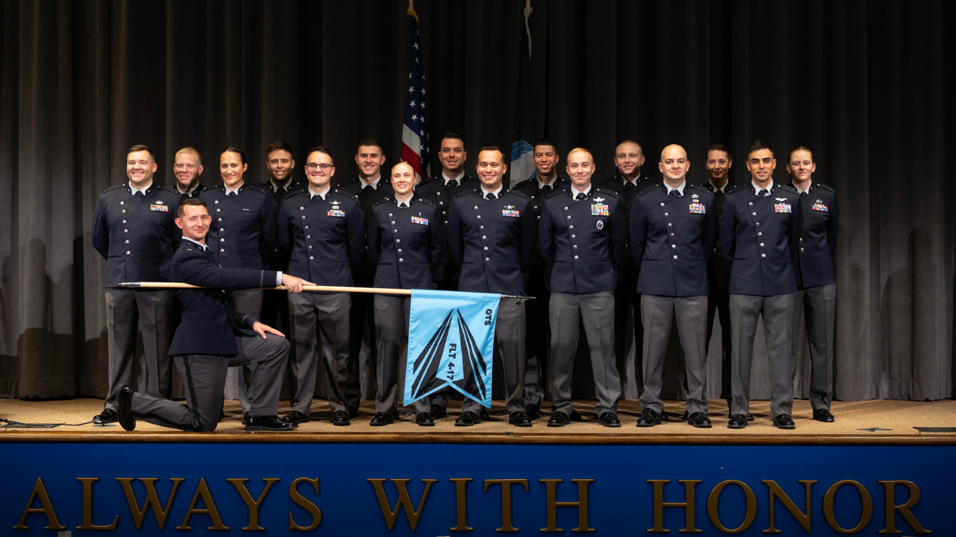 A group of people wearing dark blue shirts and black pants stand on stage with an American flag behind them and an officer kneeling down with a blue flag for the US Space Force placed in front of them.