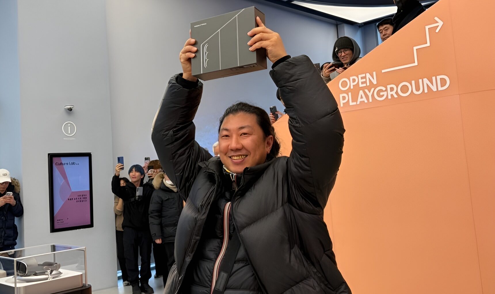 A person holding a box above their head stands near a sign that says 'OPEN PLAYGROUND,' surrounded by people taking photos with smartphones.