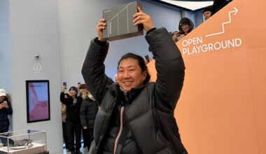 A person holding a box above their head stands near a sign that says 'OPEN PLAYGROUND,' surrounded by people taking photos with smartphones.