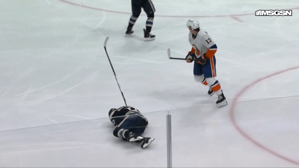 An ice hockey player in a black and white uniform lies on the ice next to a fallen stick, while another player in an orange and blue uniform skates past.