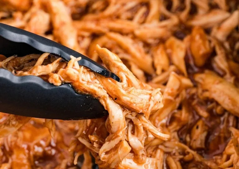 Black tongs holding shredded chicken coated in BBQ sauce, with more saucy shredded chicken in the background.