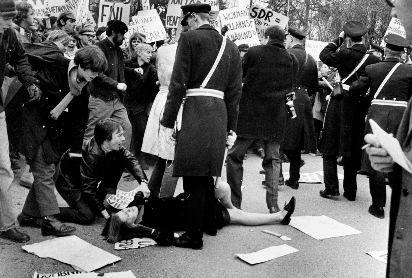 A black and white photograph of a protest in which many people are carrying signs written in Swedish, and one woman has fallen to the ground, whle another person is crouched next to her. A policeman in a long coat looks down at her