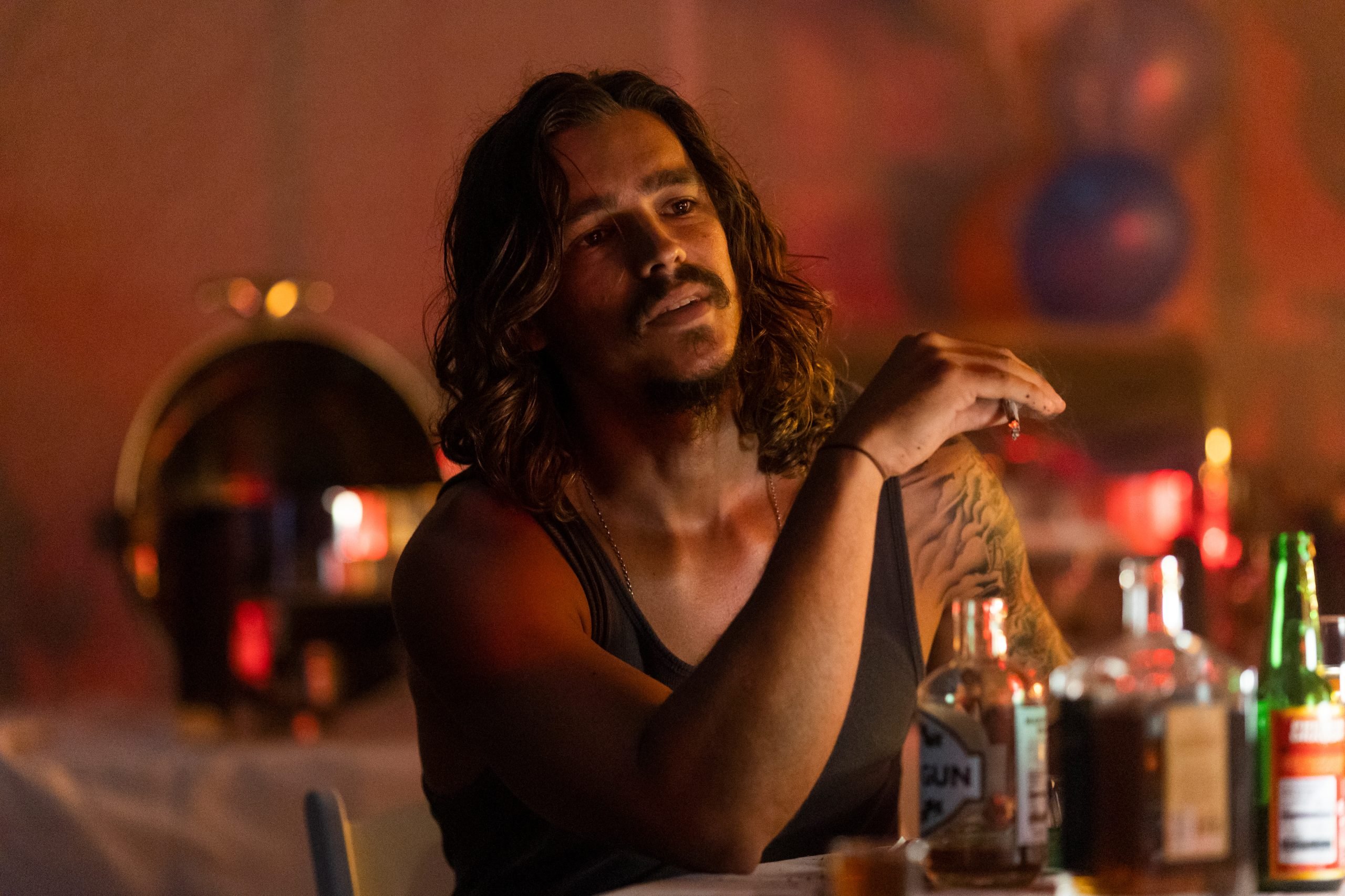 A man with long hair and a tattooed arm sits at a table with bottles, wearing a dark tank top in a dimly lit room.