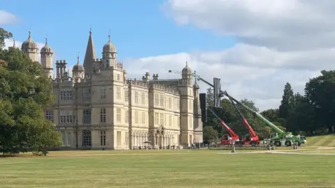 A stately home with cranes holding filming equipment in place for filming