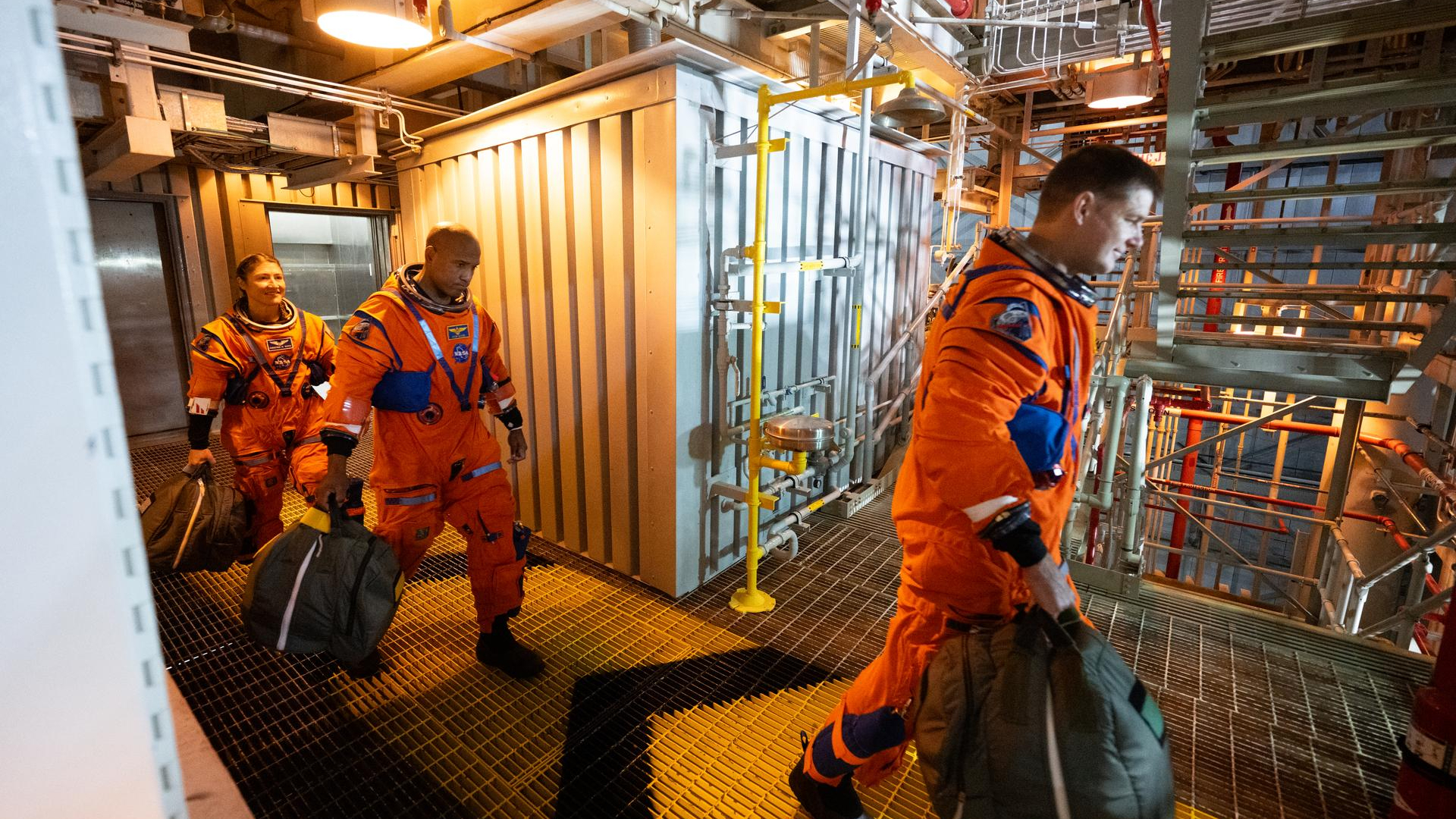 two male astronauts and a female astronaut, all wearing orange flight suits, walk on a metallic gantry inside a huge building
