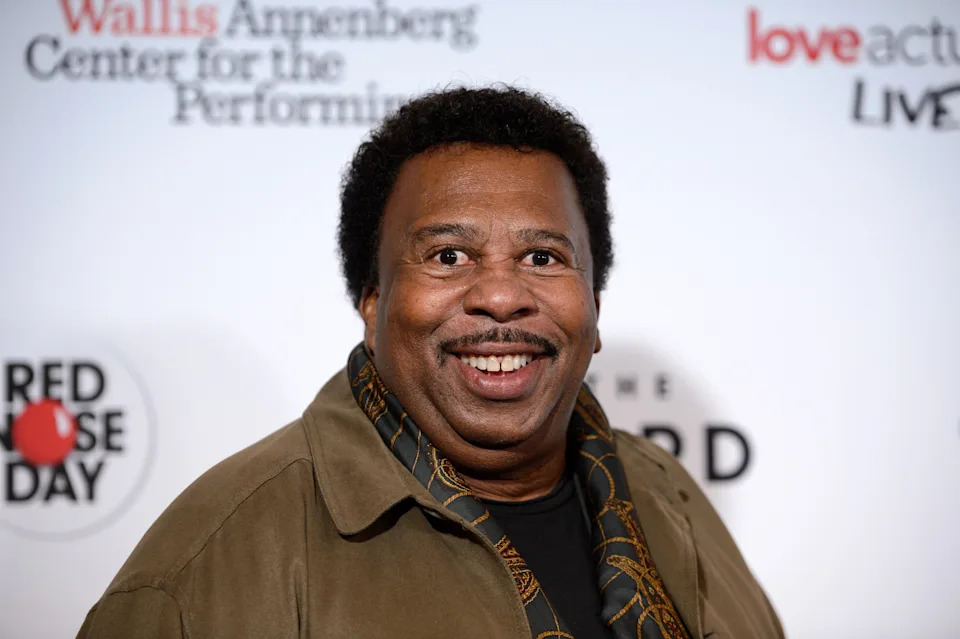 A person smiling on a red carpet, wearing a casual overcoat and patterned scarf. Event logos in the background