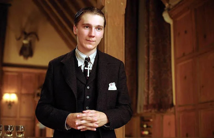 A person wearing a vintage-style suit stands indoors with hands clasped, in a scene that suggests a period drama or movie setting
