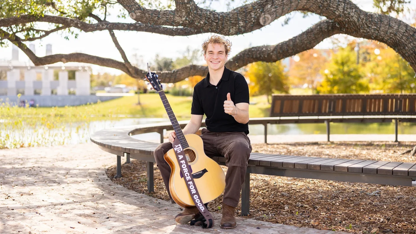 Portrait of Aiden Ross holding his acoustic guitar with a Texas A&M "Force for Good" guitar strap