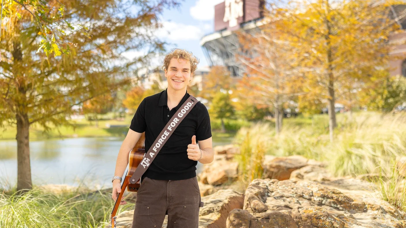 Portrait of Aiden Ross holding his acoustic guitar with a an Texas A&M "Force for Good" guitar strap in Aggie Park