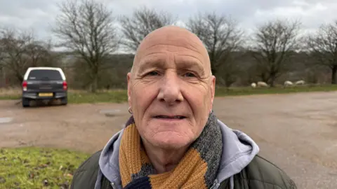 Martin Heath/BBC Paul Cox with very short hair looking at the camera and wearing a green jacket with a grey hood, and a yellow scarf. He is standing in a car park with one car in a space behind him and trees beyond. There are also four sheep in the background.
