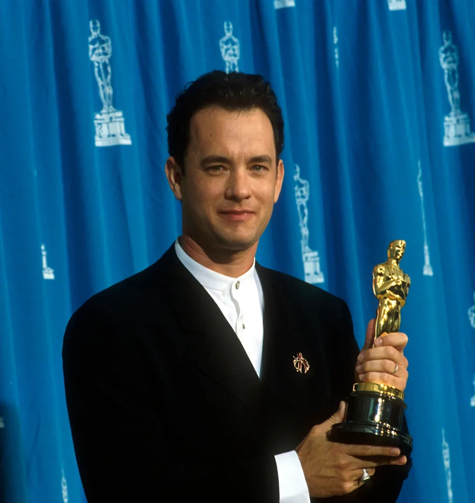 Person holding an Oscar trophy, wearing a black suit with a white shirt, stands in front of a backdrop featuring Oscar statues
