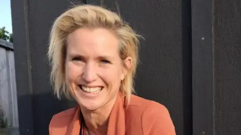 Kim Black A woman smiling with blonde hair wearing an orange blazer. 