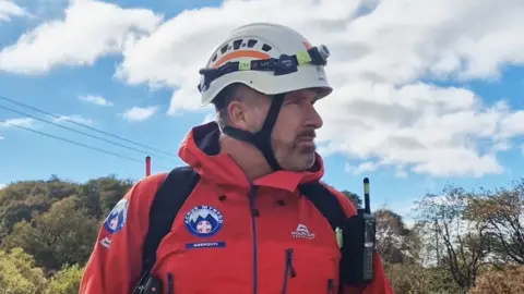 Gareth Davies Gareth Davies is a middle-aged man with short greying hair and a closely trimmed beard and mustache. His head and torso is visible and he is looking to the right of the picture. He wears a white climbing safety helmet with straps and an integrated head torch, and a bright orange outdoor jacket with a white and blue mountain rescue logo on the chest and arm. He is carrying a backpack which has a walkie-talkie in a waterproof cover attached. In the background are blue sky with clouds and trees.