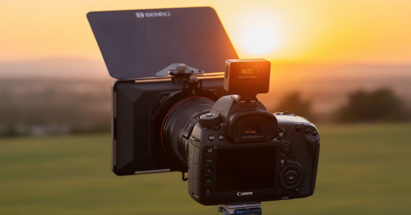 A Canon DSLR camera mounted on a tripod faces a sunset, equipped with a large ND filter and hood. The background is a blurred landscape of green fields and a glowing orange sky.