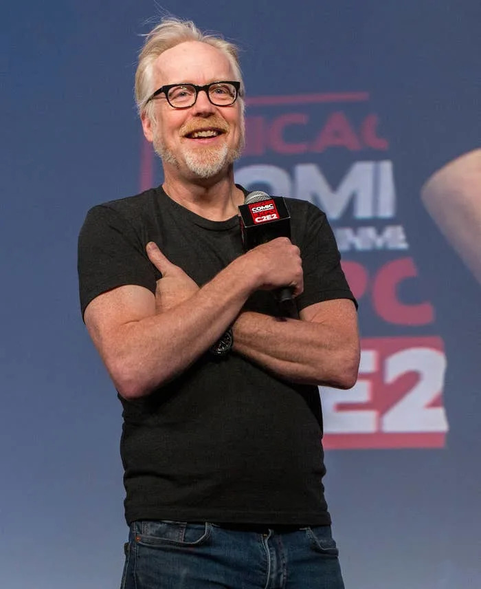 A person with glasses and a beard stands on stage at a comic convention, smiling while holding a microphone