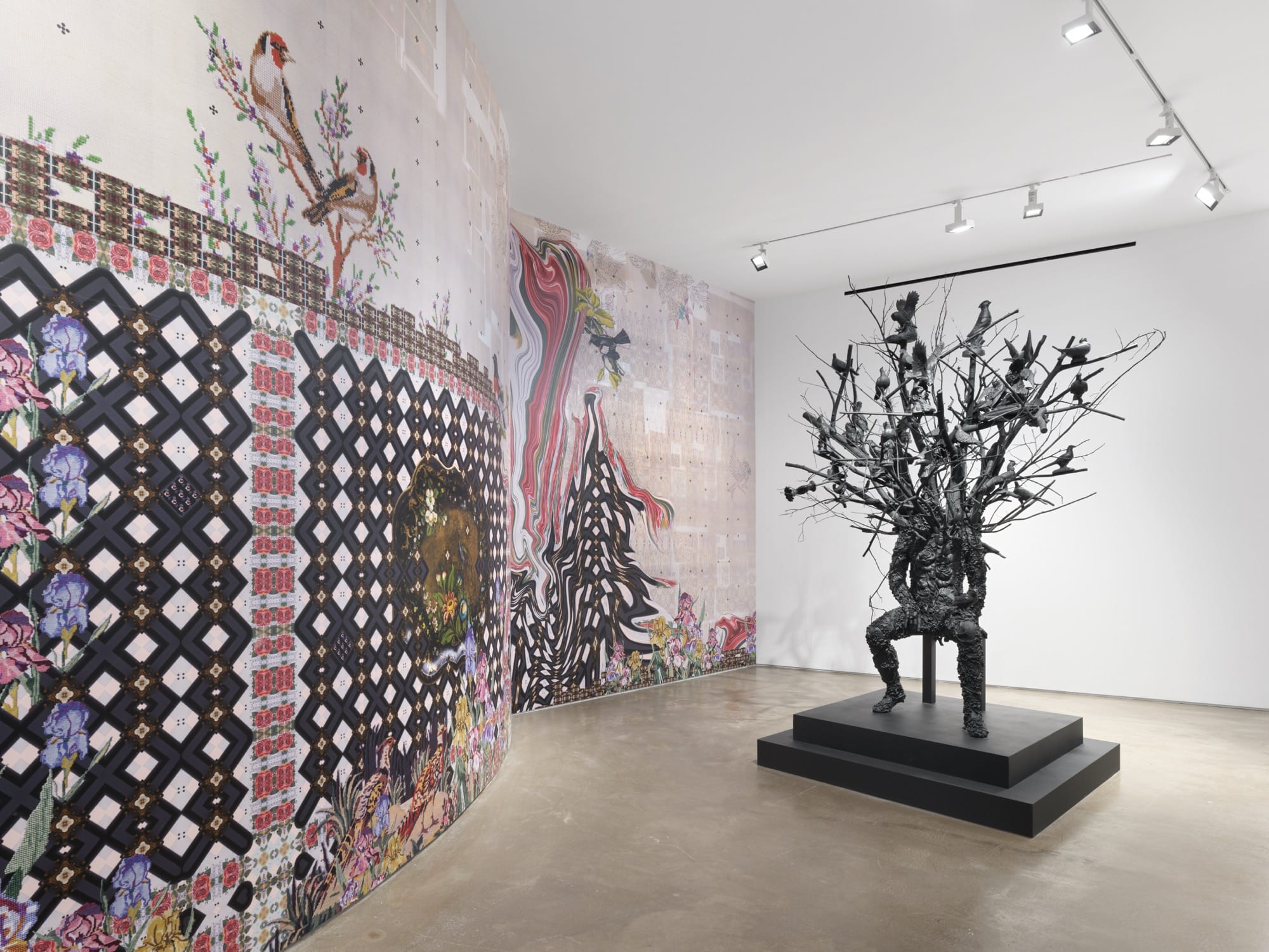 a large-scale bronze figure with branches and leaves for a head sits on a chair next to elaborate wallpaper