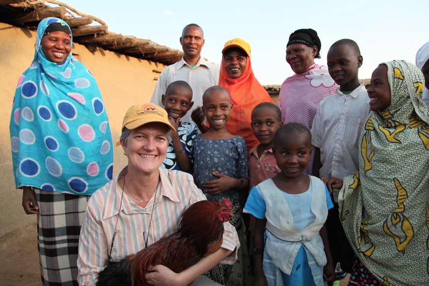 A woman holiding a chicken in her arms in a village in Tanzania with a group of villagers in traditional dress looking on