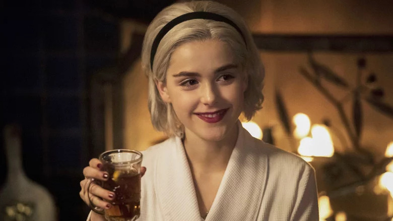 Sabrina Spellman (Kiernan Shipka) grins while holding a cup of tea by a fire in Chilling Adventures of Sabrina