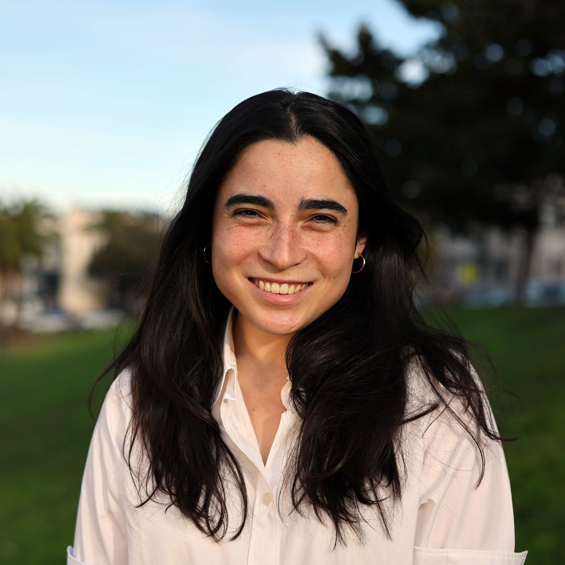 Person with long dark hair smiles outdoors, wearing a light-colored shirt.
