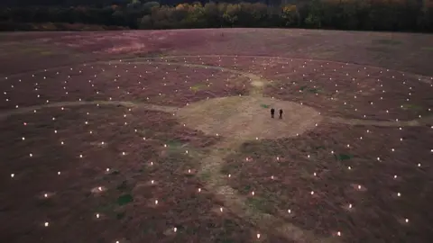 Avon Needs Trees/Elastic Pie Films A drone image of two people dressed in dark clothing, who are seen from very far away in a clearing. Around them is a large swirling artwork, lit by hundreds of lanterns spreading out in every direction. There are curved paths running through the installation. 