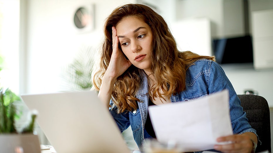 A worried woman sits at a table reviewing her household budget and bills, appearing stressed about home finances.