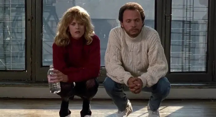 Two people kneel indoors by large windows. The woman holds a water bottle and wears a sweater; the man wears a cable-knit sweater and sneakers