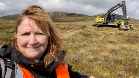 PA Selfie-style picture of Lorna Dawson, who has long brown hair. She is wearing a black jacket and and an orange hi-viz vest. Moorland and hills can be see in the background and a large digger is visible in the background.