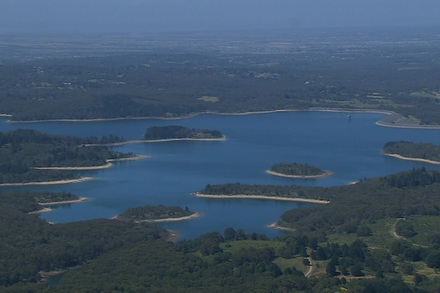 An aerial shot of a large dam surrounded by green trees with islands covered in green trees in the water.